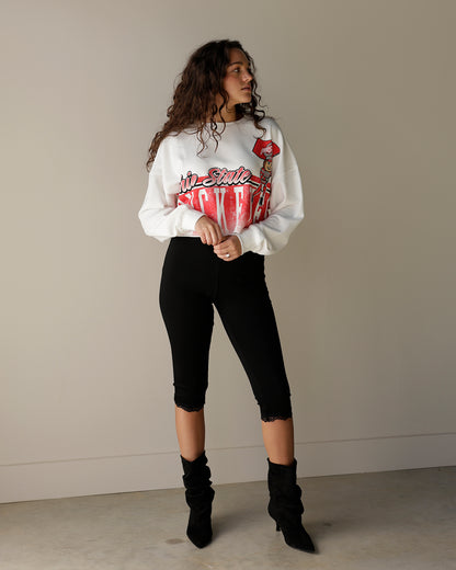 A woman modeling a white crewneck sweatshirt with a red and black graphic print, paired with black cropped leggings and black ankle boots, standing against a plain beige wall.