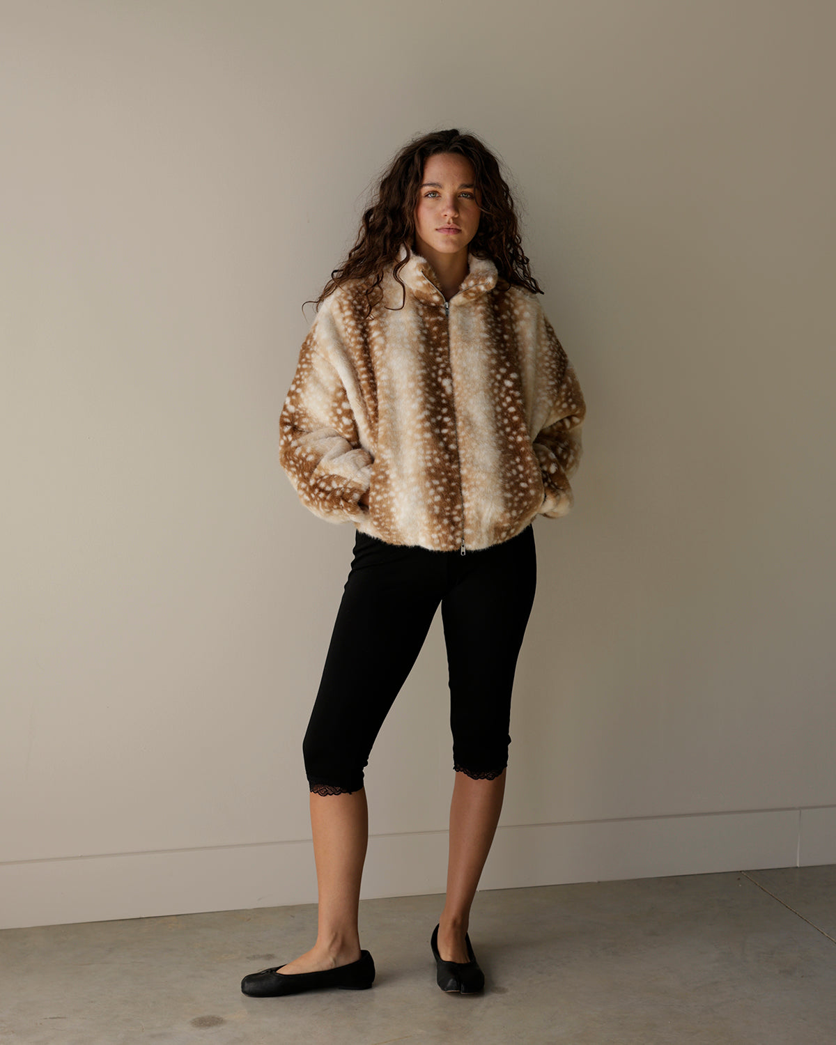 A woman stands against a plain beige wall wearing a fuzzy cropped jacket with a brown and cream spotted pattern, black cropped pants, and black flats.