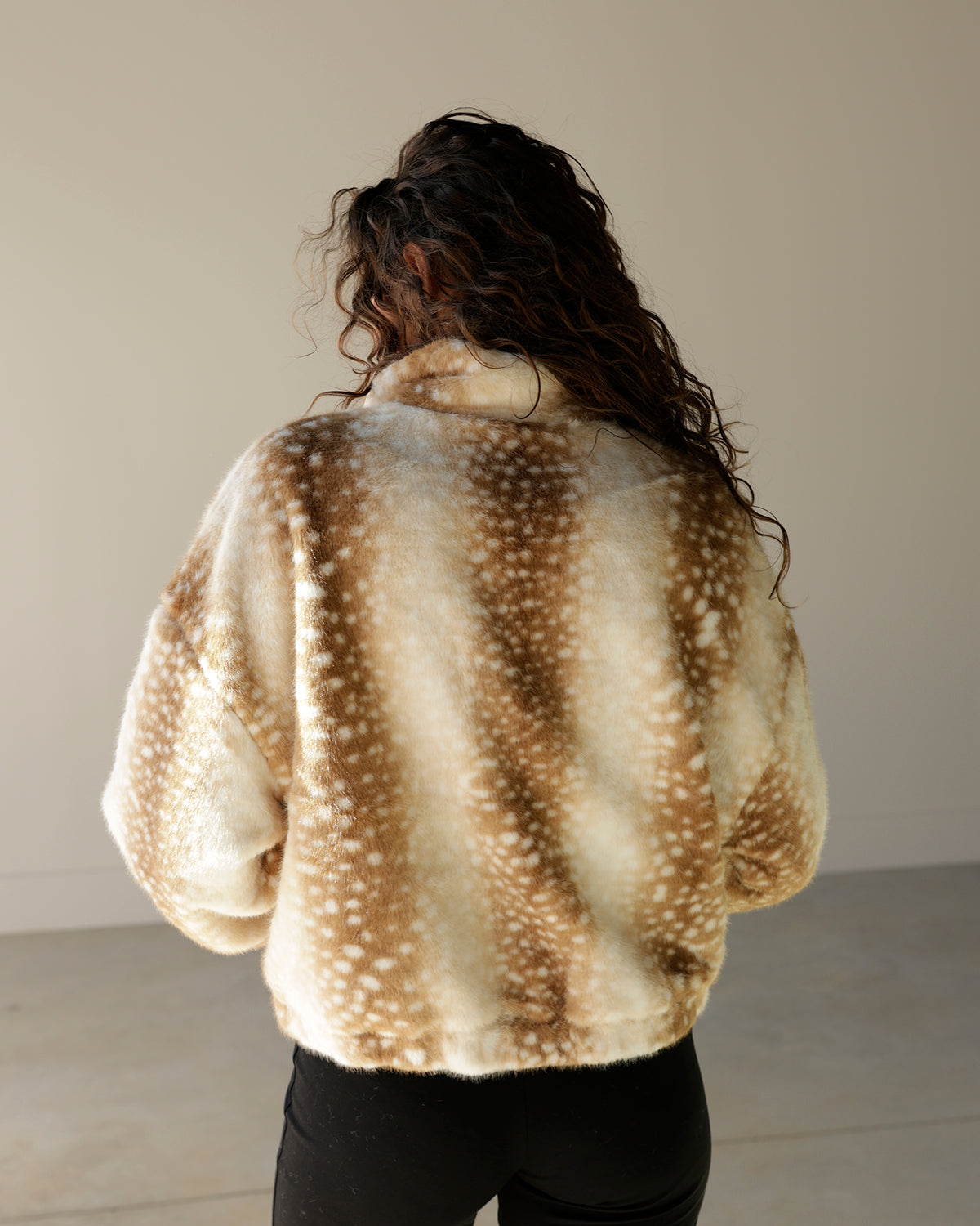 A person viewed from behind wearing a fuzzy cropped jacket with a brown and cream mottled animal-inspired spot pattern, paired with black pants, against a plain indoor wall.