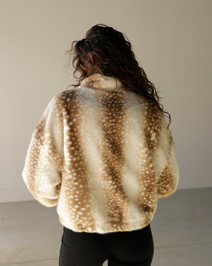 A person viewed from behind wearing a fuzzy cropped jacket with a brown and cream mottled animal-inspired spot pattern, paired with black pants, against a plain indoor wall.