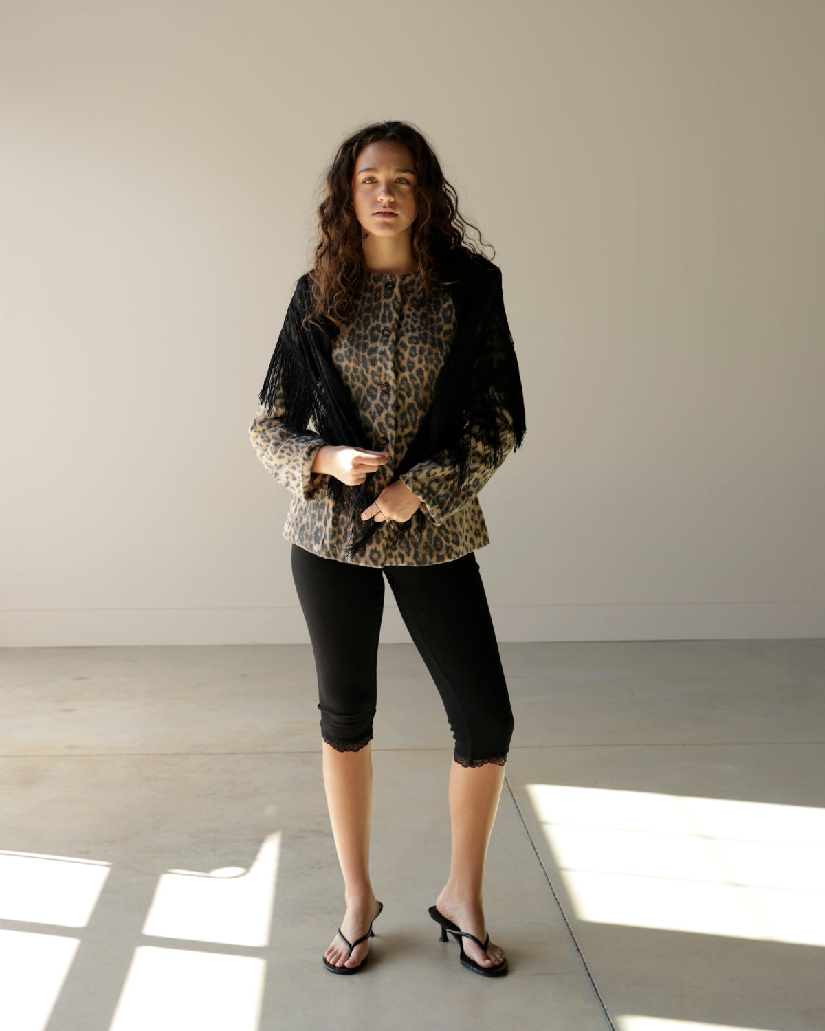 A woman modeling a black sheer mesh jacket over a leopard print top, paired with black capri leggings and black thong sandals, standing in a minimalist studio with concrete floors and soft window light.