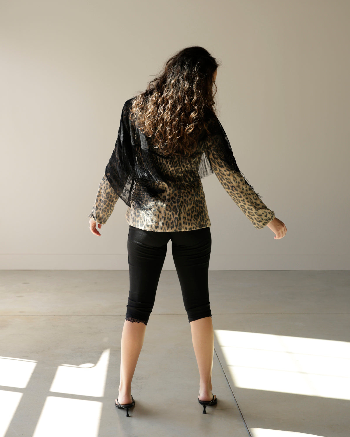 A woman stands with her back to the camera in a minimalist studio, wearing a leopard print long-sleeve top with a sheer black overlay, black capri pants, and black heels.