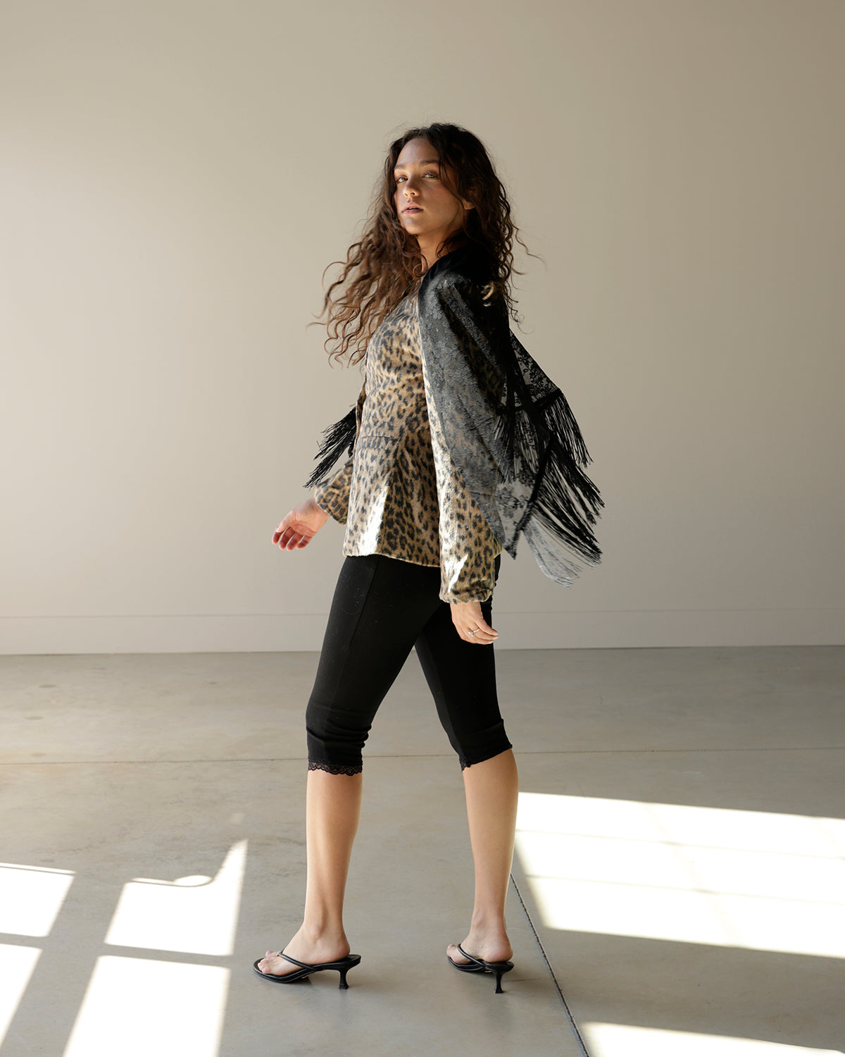 A woman models a leopard-print long-sleeve top with a black fringed shawl draped over her shoulders, paired with black capri leggings and black slingback heels, in a minimalist studio setting.