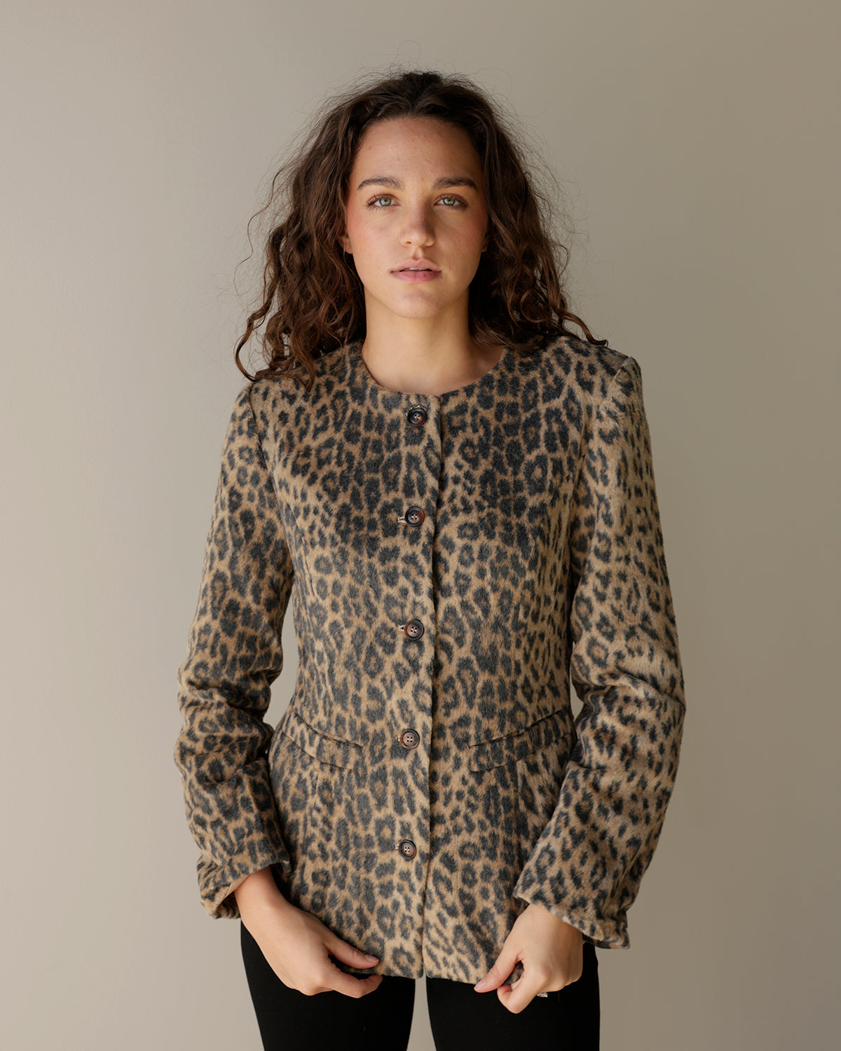 A woman modeling a leopard print button-up jacket with structured shoulders and front pockets, set against a plain warm gray studio background.