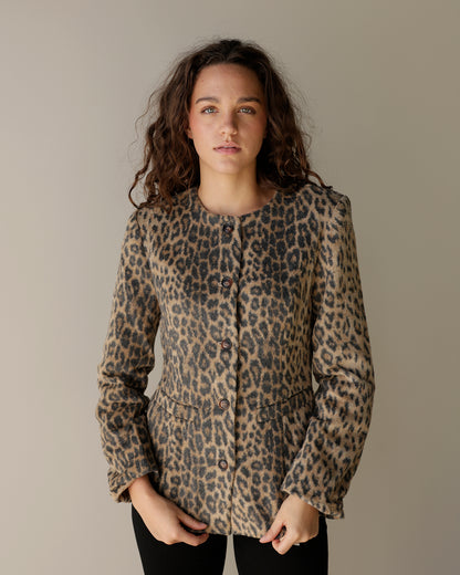A woman modeling a leopard print button-up jacket with structured shoulders and front pockets, set against a plain warm gray studio background.