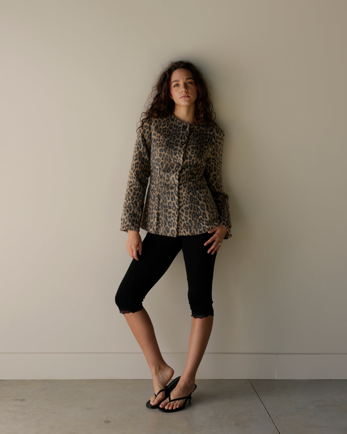 A woman stands against a plain beige wall wearing a leopard-print cropped jacket, black cropped pants, and black sandals.