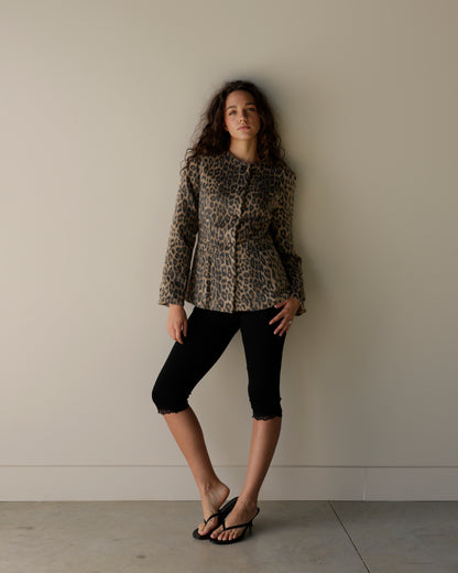 A woman stands against a plain beige wall wearing a leopard-print cropped jacket, black cropped pants, and black sandals.