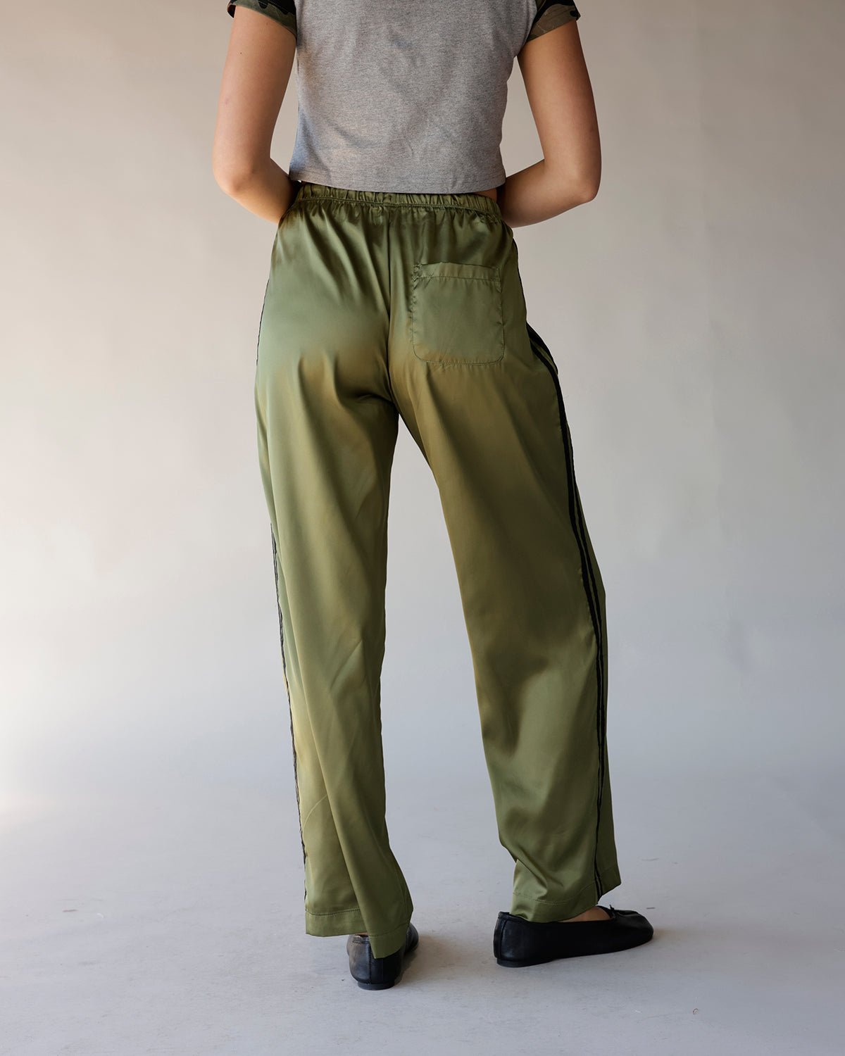 Olive green satin track pants with black side stripe and elastic waistband, back view