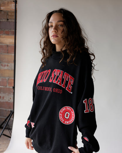Oversized black collegiate crewneck sweatshirt with red lettering and embroidered patches