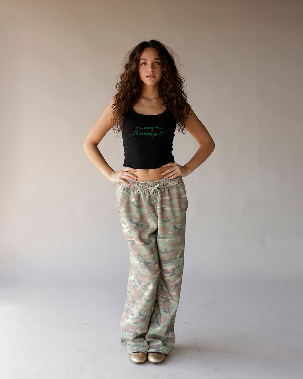 Black cropped tank top with green script graphic, styled with camo wide-leg sweatpants