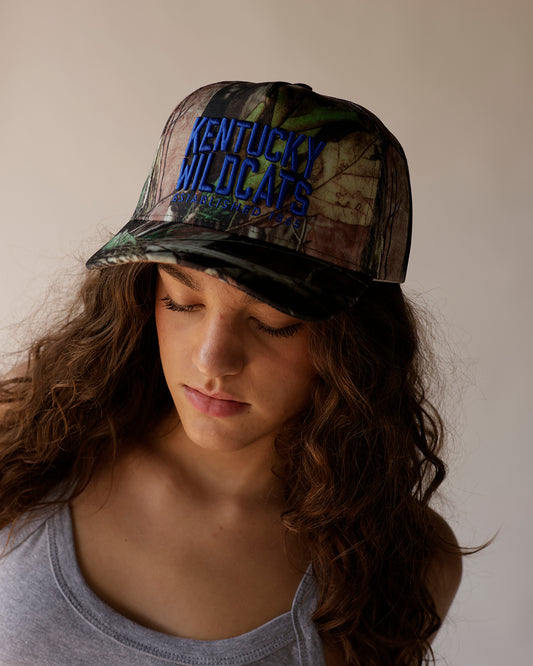 Kentucky Wildcats camo trucker hat with blue embroidered text, worn by model