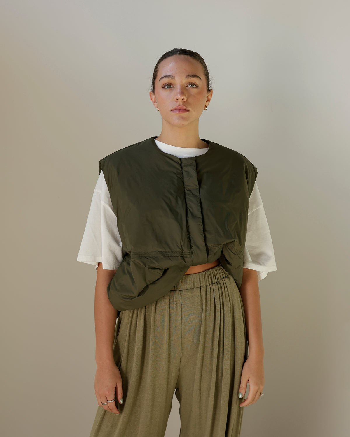 women wearing olive green vest with oversized white tshirt underneath she is wearing pleated balloon pants with her hair pulled back 