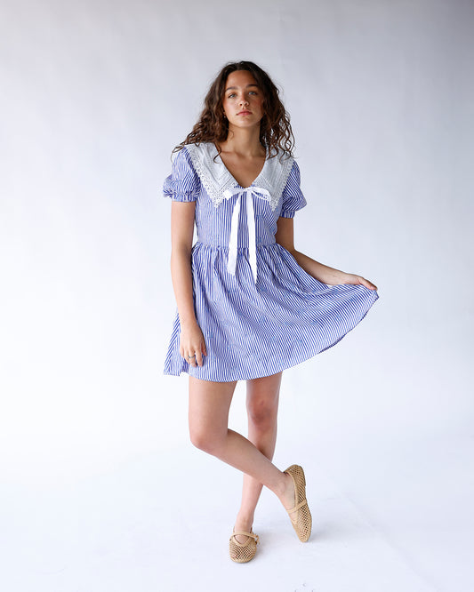 woman wearing a blue and white vertical stripe mini dress with blue shell embroidered all over and a peter pan white lace collar with front white bow to tie