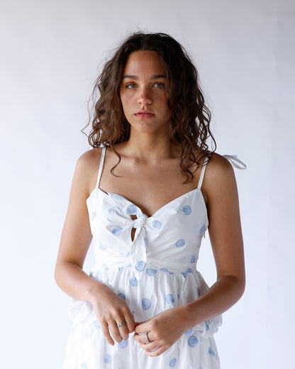 White peplum cami featuring blue embroidered seashells, spaghetti straps, a flattering v-neckline, and a front tie bow. The perfect coastal-inspired top for summer styling.
