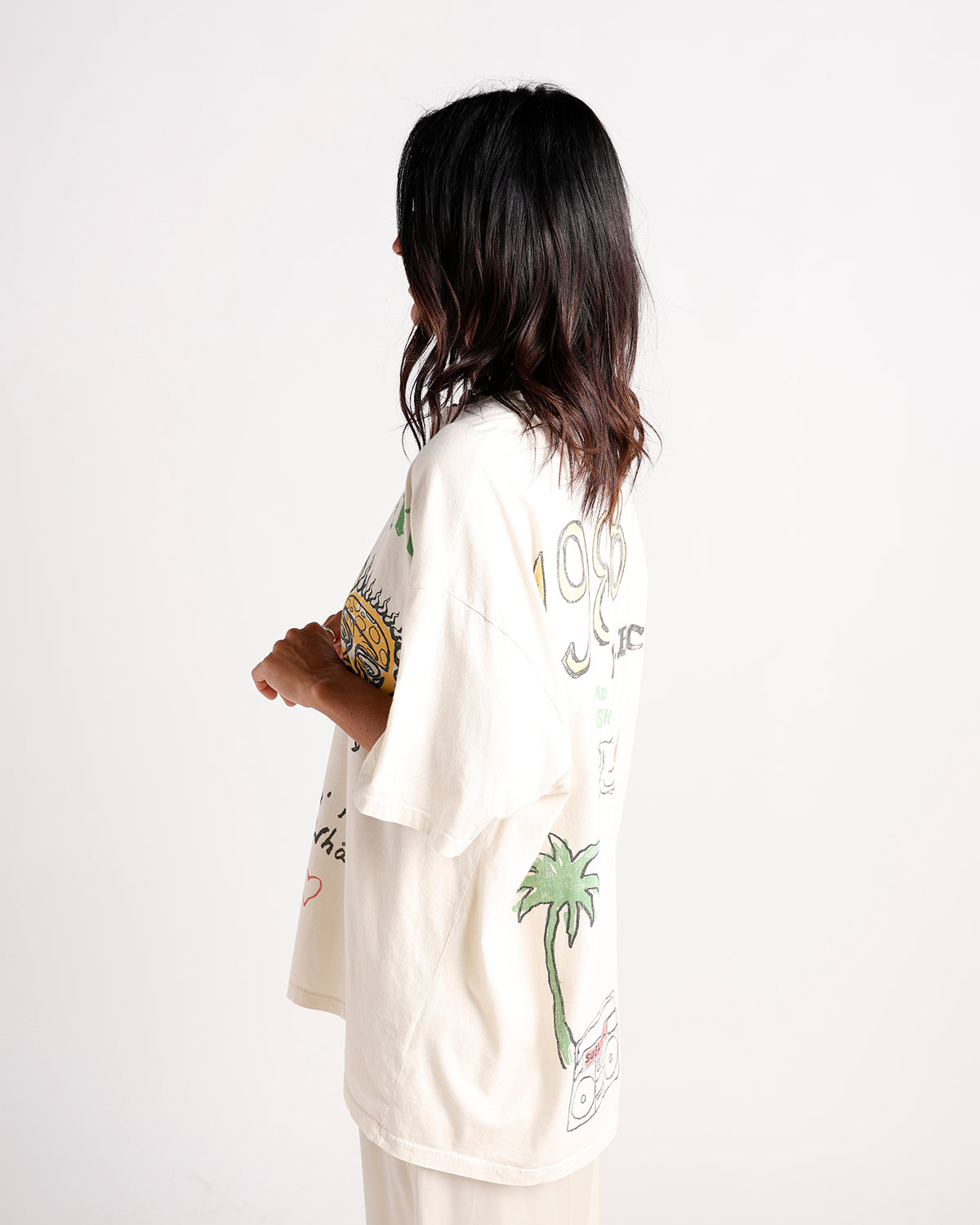 Woman wearing oversized cream graphic tee with palm tree and sun illustrations, side view