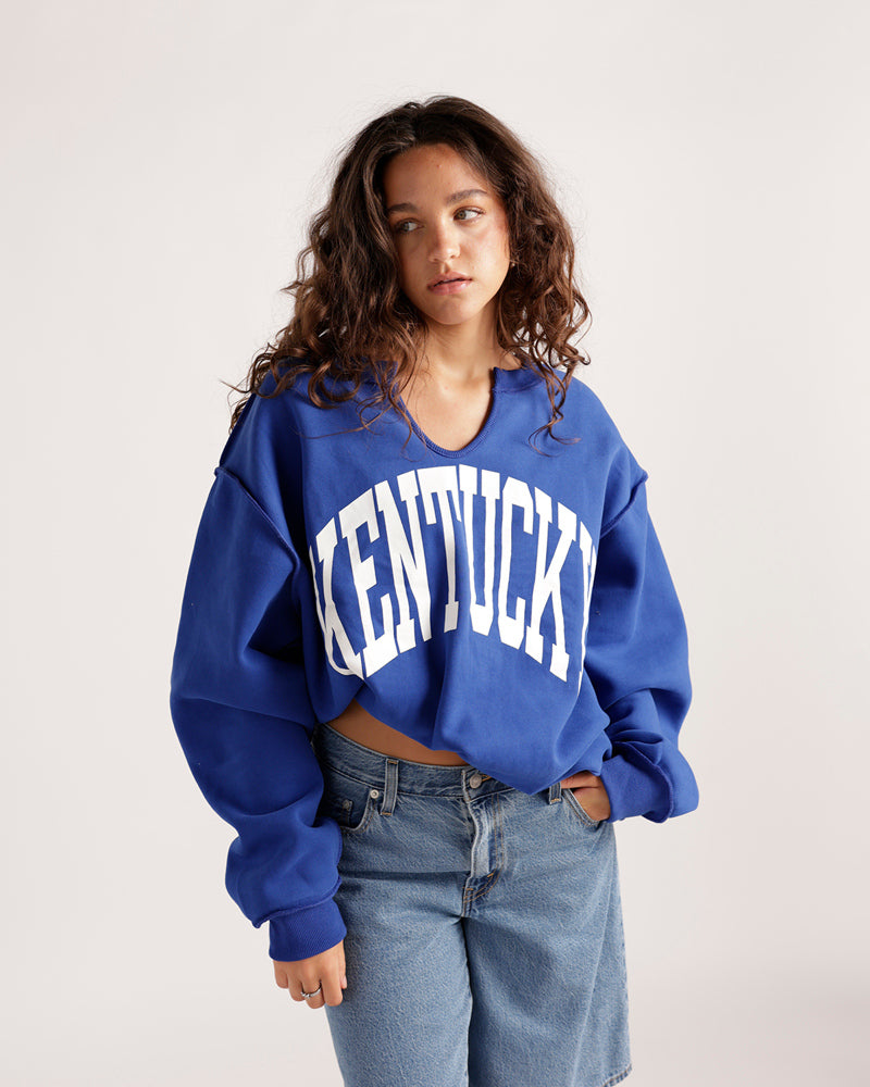 Cropped blue Kentucky sweatshirt with V-neck and white arched lettering, worn with jeans
