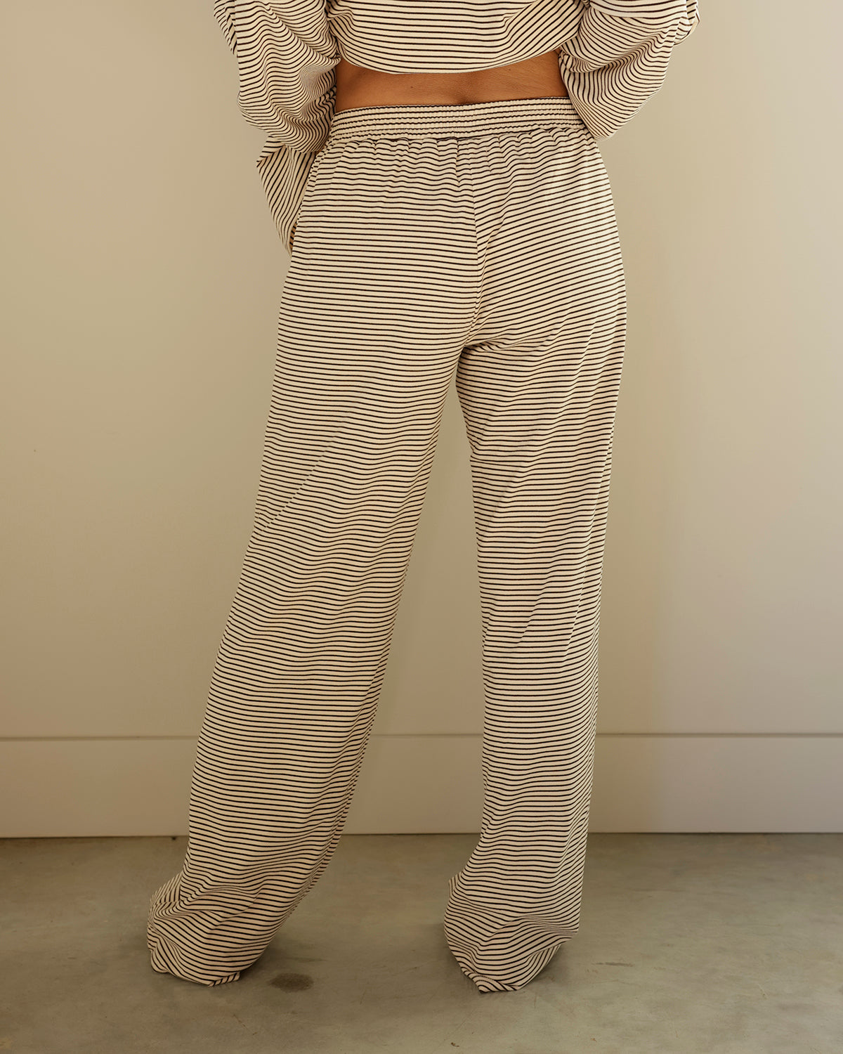 A person shown from the back wearing loose, wide-leg pants and a matching striped crop top in beige and white, standing against a plain beige wall and concrete floor.