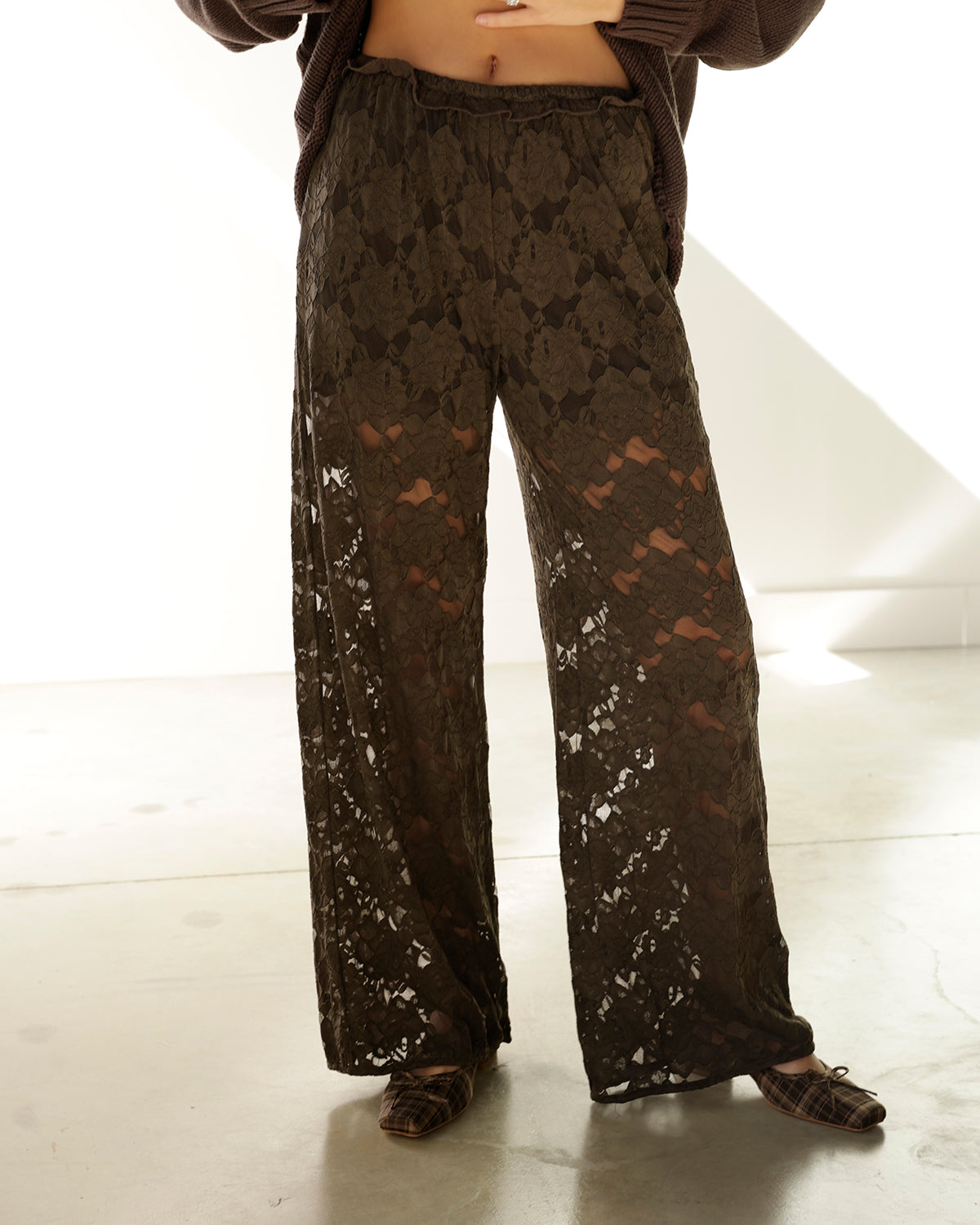 A pair of dark gray lace pants hanging on a rack, with a hand holding the hem to show the drape. A white tag with the brand name 'aren' is visible on the right side.