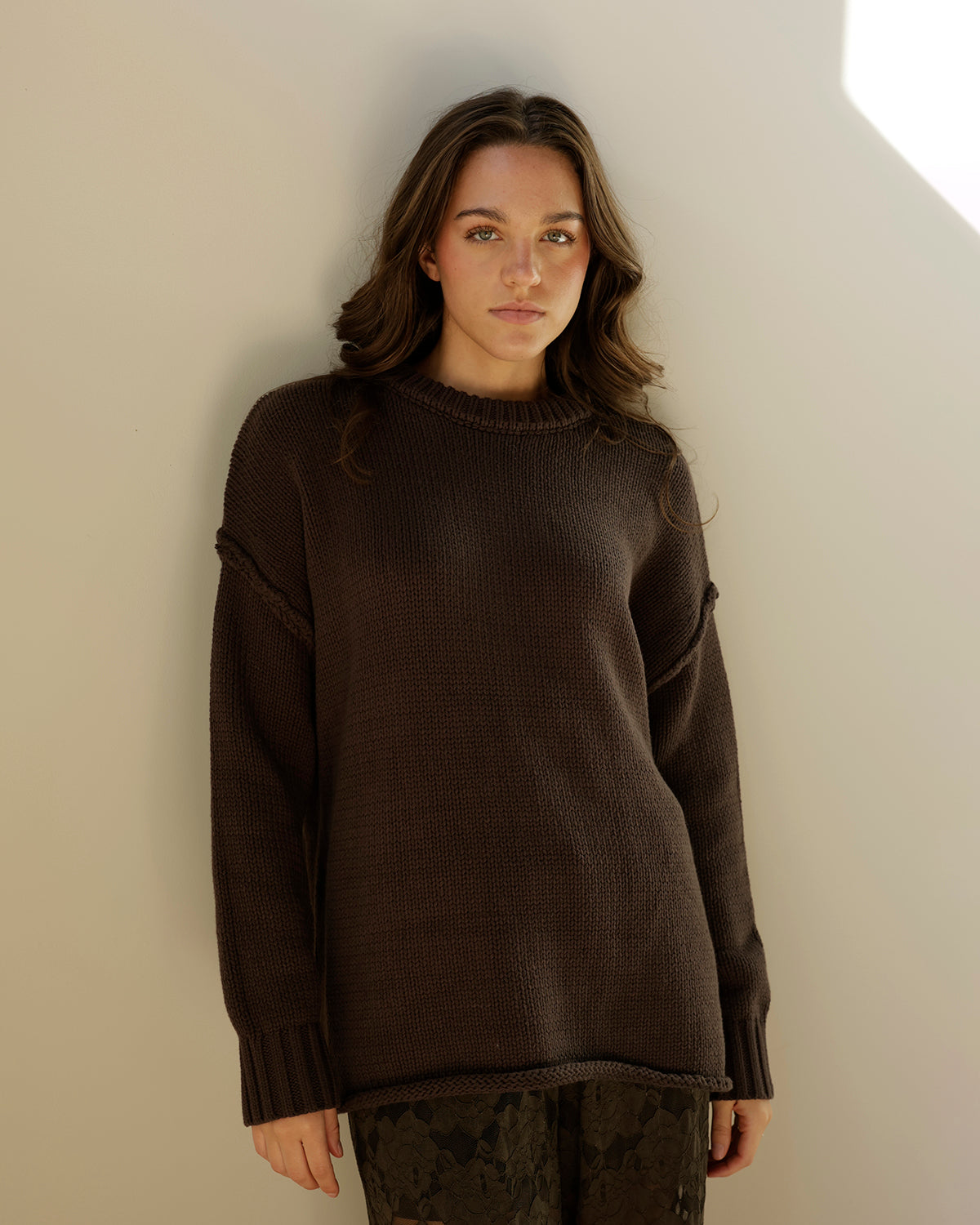 A dark brown ribbed knit sweater hanging on a wooden hanger, shown next to other sweaters on a clothing rack.