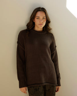 A dark brown ribbed knit sweater hanging on a wooden hanger, shown next to other sweaters on a clothing rack.