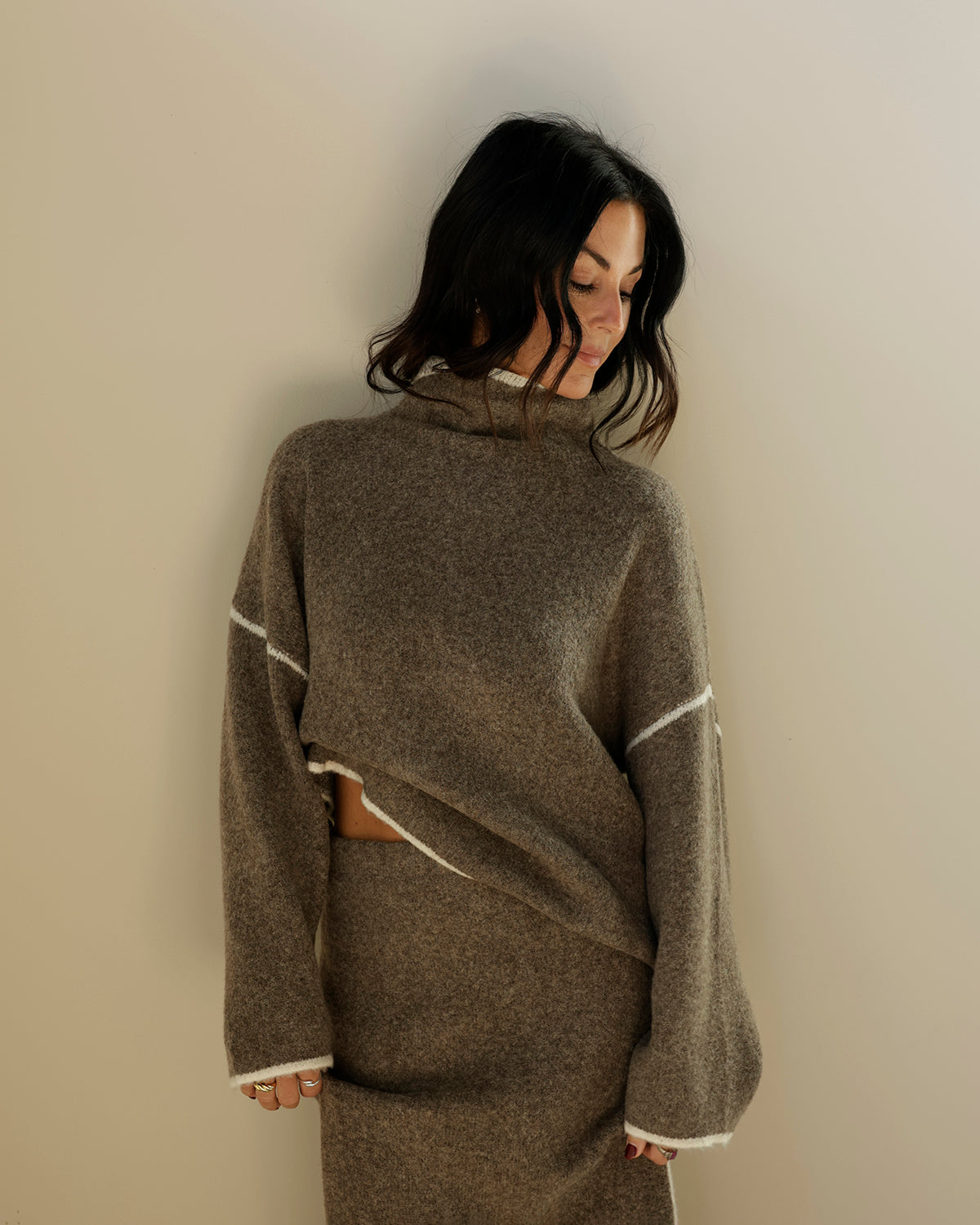 A person's hand holding up a gray wool turtleneck poncho sweater with white contrast stitching, photographed in a retail store setting.