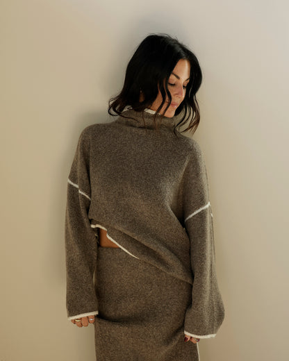 A person's hand holding up a gray wool turtleneck poncho sweater with white contrast stitching, photographed in a retail store setting.