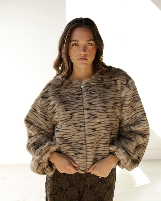 A woman wearing a cropped faux fur jacket with a brown and beige tiger-stripe pattern, standing against a bright white background.