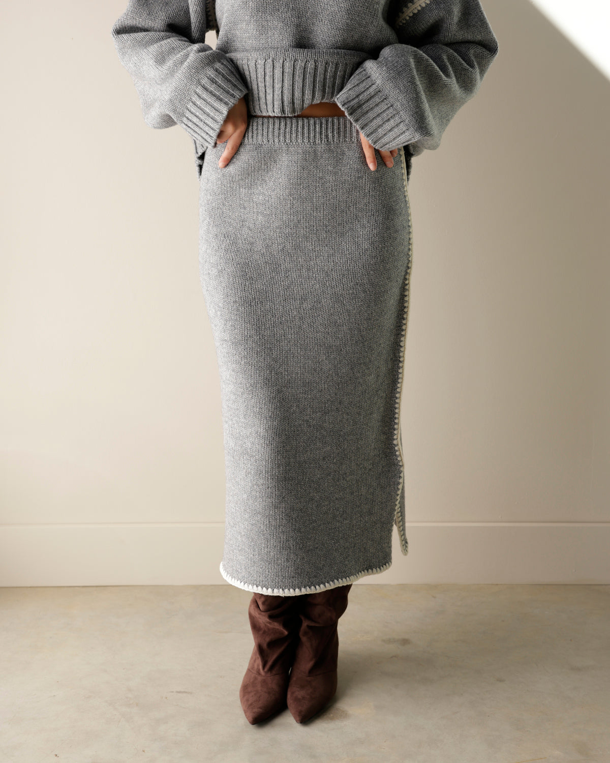 A close-up of a gray knit sweater skirt hanging on a hanger, with a hand holding the edge to show the white zigzag trim. A product tag is attached, and a wooden hanger and wood background are visible.