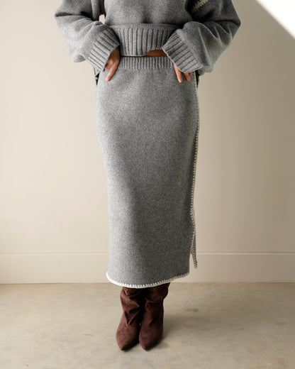 A close-up of a gray knit sweater skirt hanging on a hanger, with a hand holding the edge to show the white zigzag trim. A product tag is attached, and a wooden hanger and wood background are visible.