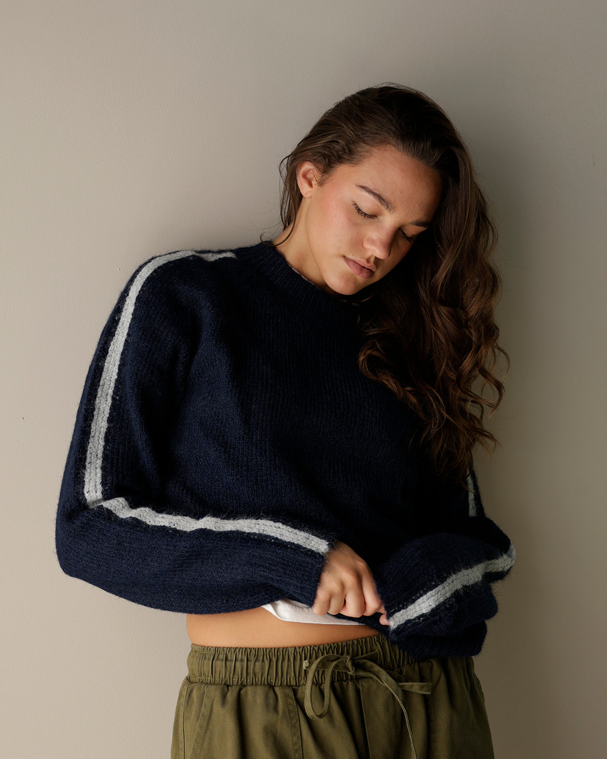A fuzzy knit crewneck sweater in light oatmeal beige hanging on a wooden hanger, with navy and brown striped sweaters visible behind it on a clothing rack.