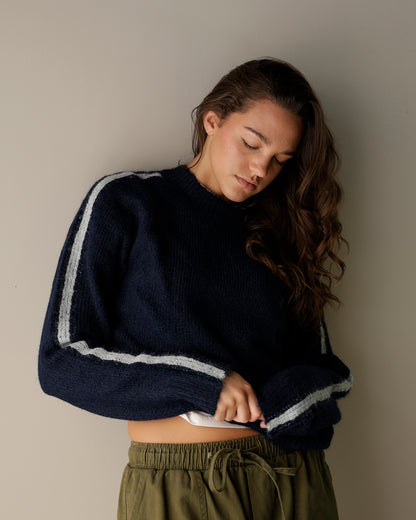 A fuzzy knit crewneck sweater in light oatmeal beige hanging on a wooden hanger, with navy and brown striped sweaters visible behind it on a clothing rack.