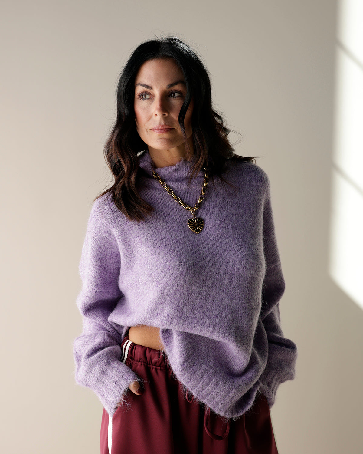 A lavender purple knit mock-neck sweater hanging on a wooden hanger, with other garments partially visible on the sides.