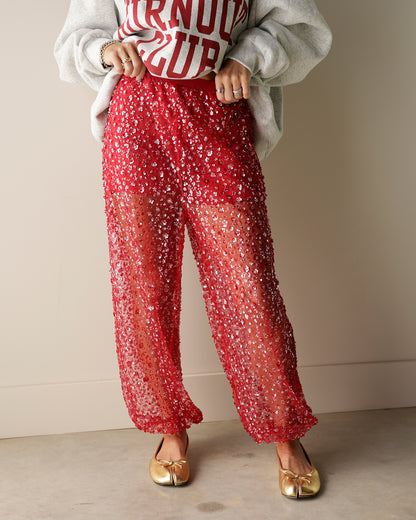 A person wearing sheer red wide-leg pants covered in small white floral sequins, styled with a white sweatshirt and gold flats against a plain beige wall.