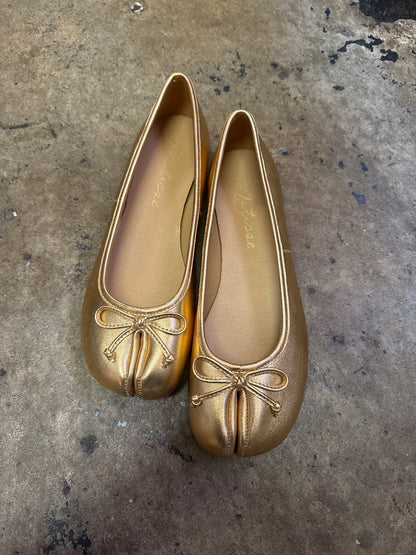 Gold metallic split-toe ballet flats with bow detail on textured concrete surface