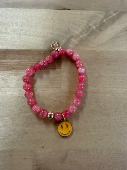 Pink beaded stretch bracelet with yellow enamel smiley face charm on wood surface