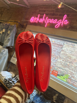 Red split-toe ballet flats with bow detail, shown sole-up in a boutique setting.