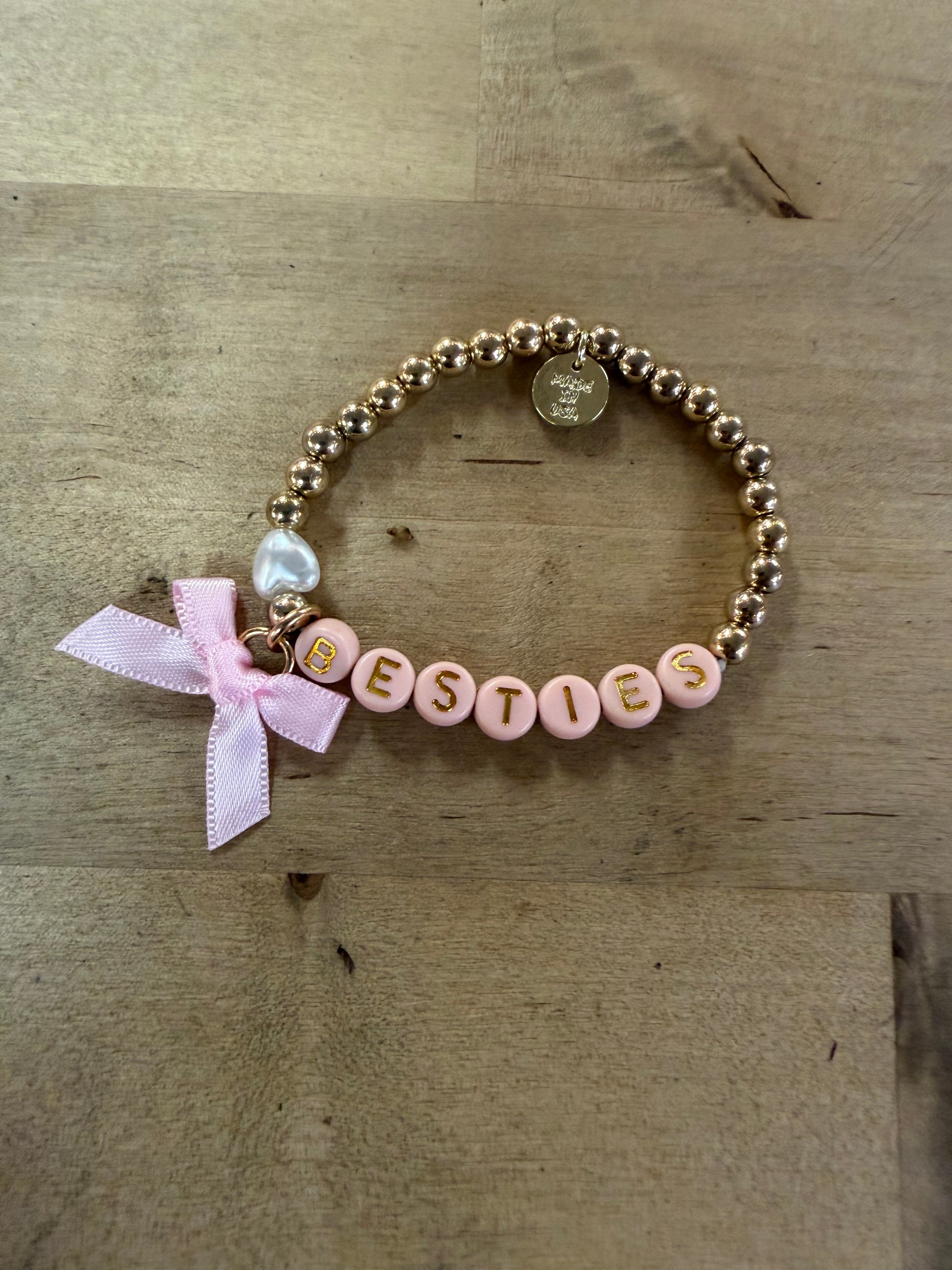 Gold beaded stretch bracelet spelling BESTIES with pink letter beads and pink bow charm