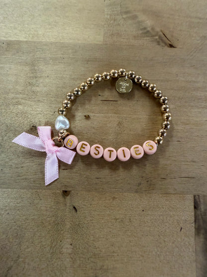 Gold beaded stretch bracelet spelling BESTIES with pink letter beads and pink bow charm