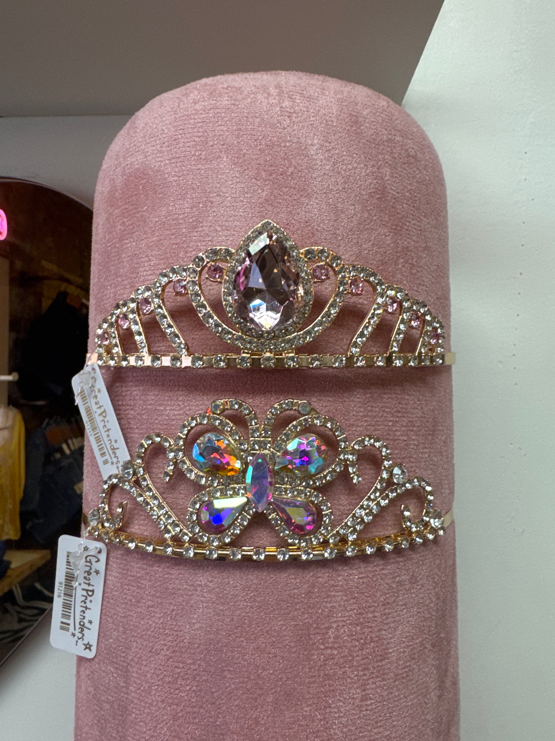 Two gold rhinestone tiaras with colorful gemstone centers displayed on pink velvet stand
