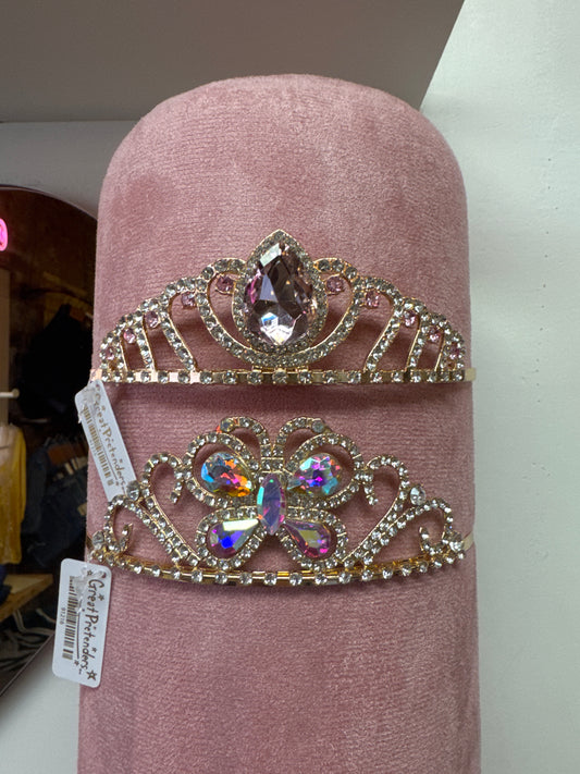 Two gold rhinestone tiaras with colorful gemstone centers displayed on pink velvet stand