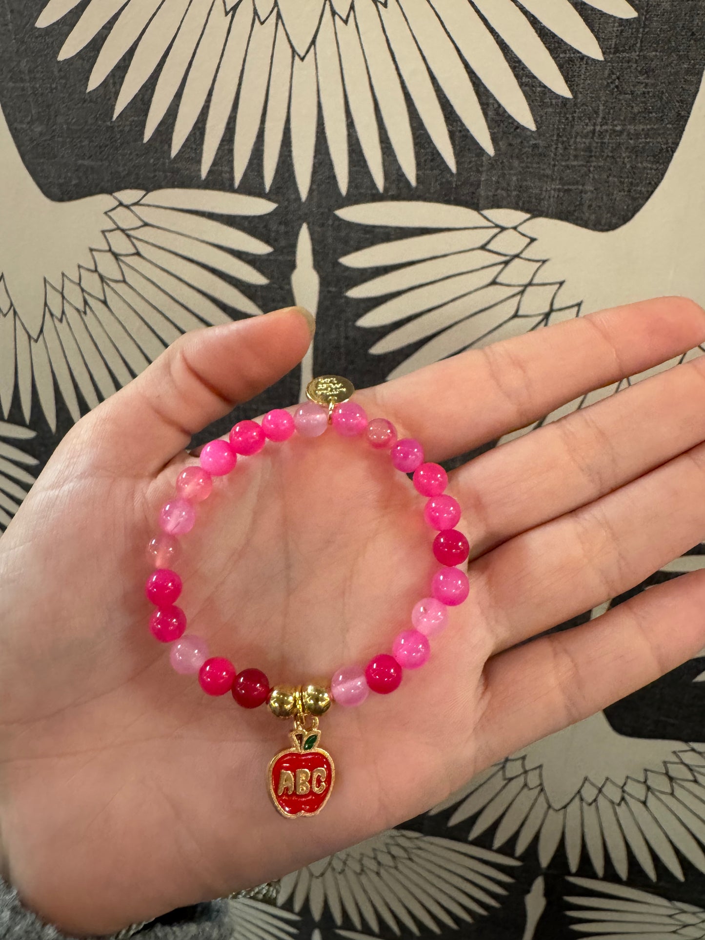 Pink beaded stretch bracelet with red enamel ABC apple charm held in palm