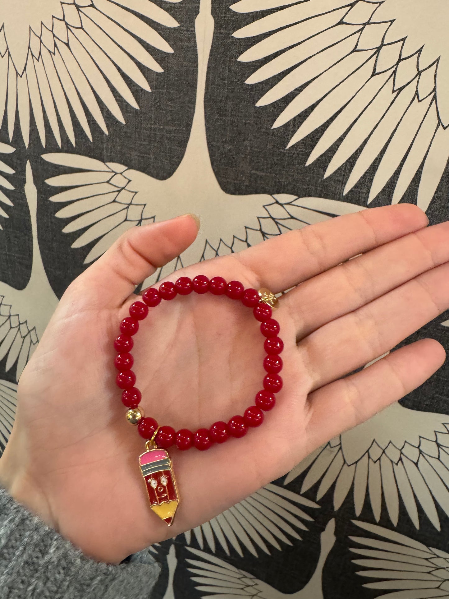 Red beaded stretch bracelet with gold-toned enamel pencil charm, held in palm