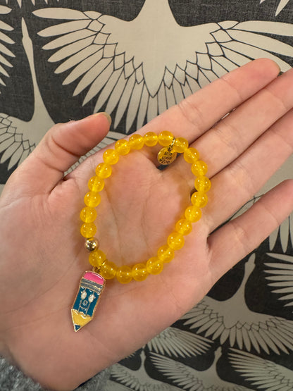 Yellow beaded stretch bracelet with colorful enamel pencil charm, held in palm