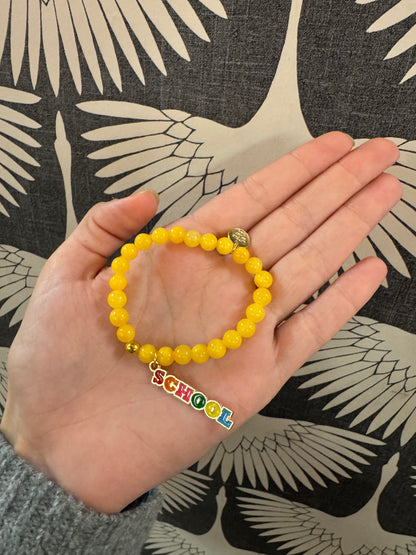 Yellow beaded stretch bracelet with colorful enamel SCHOOL charm, held in palm
