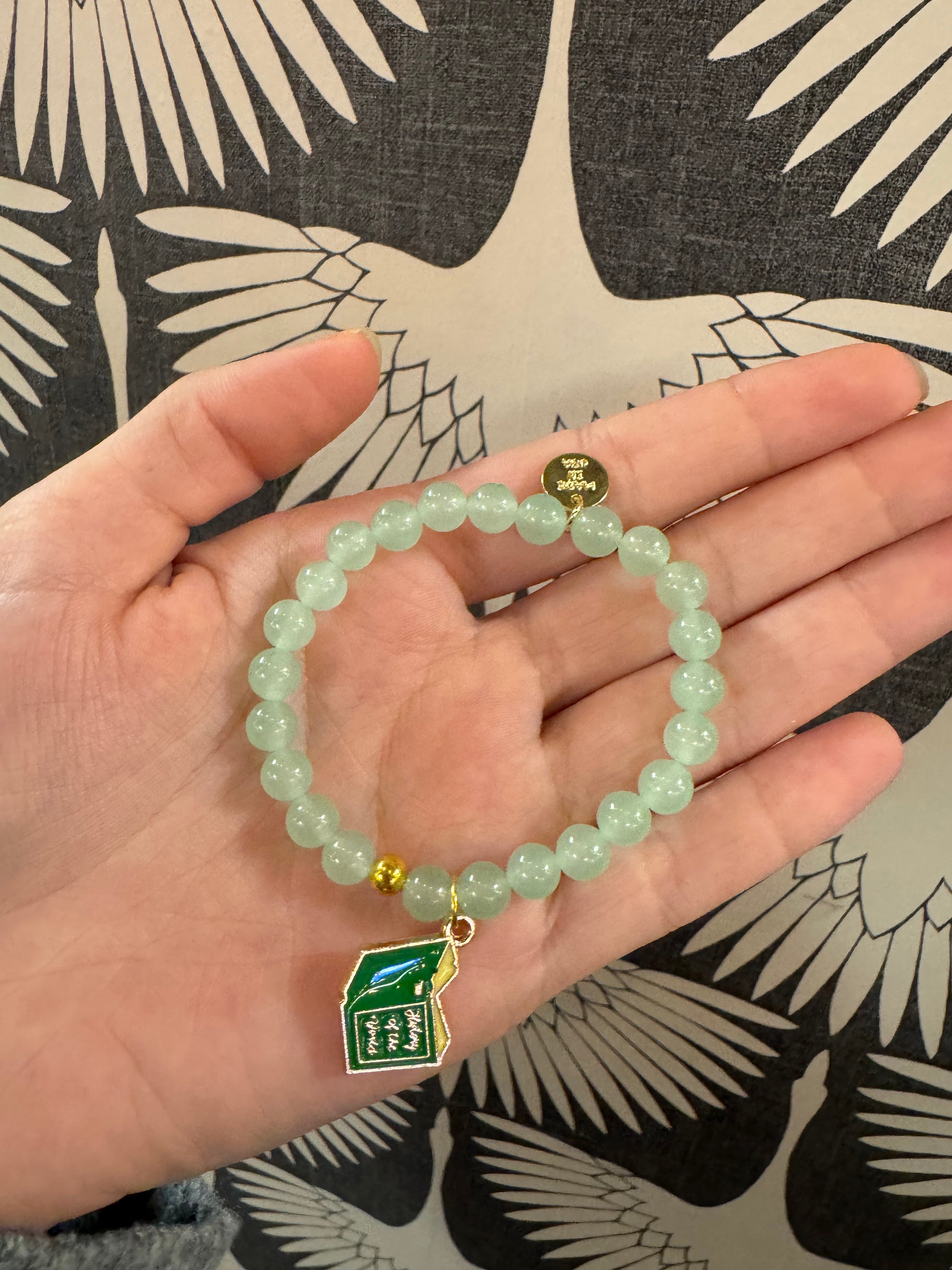 Light green beaded bracelet with enamel book charm held in open palm