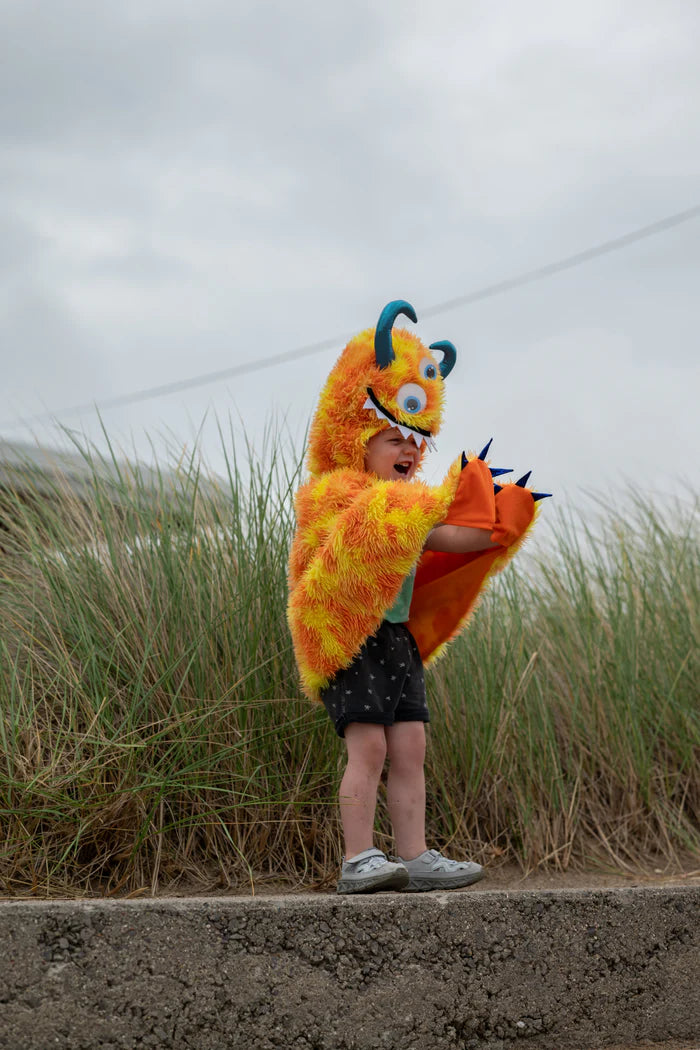 Child wearing orange fuzzy monster cape with googly eyes and teal horns outdoors