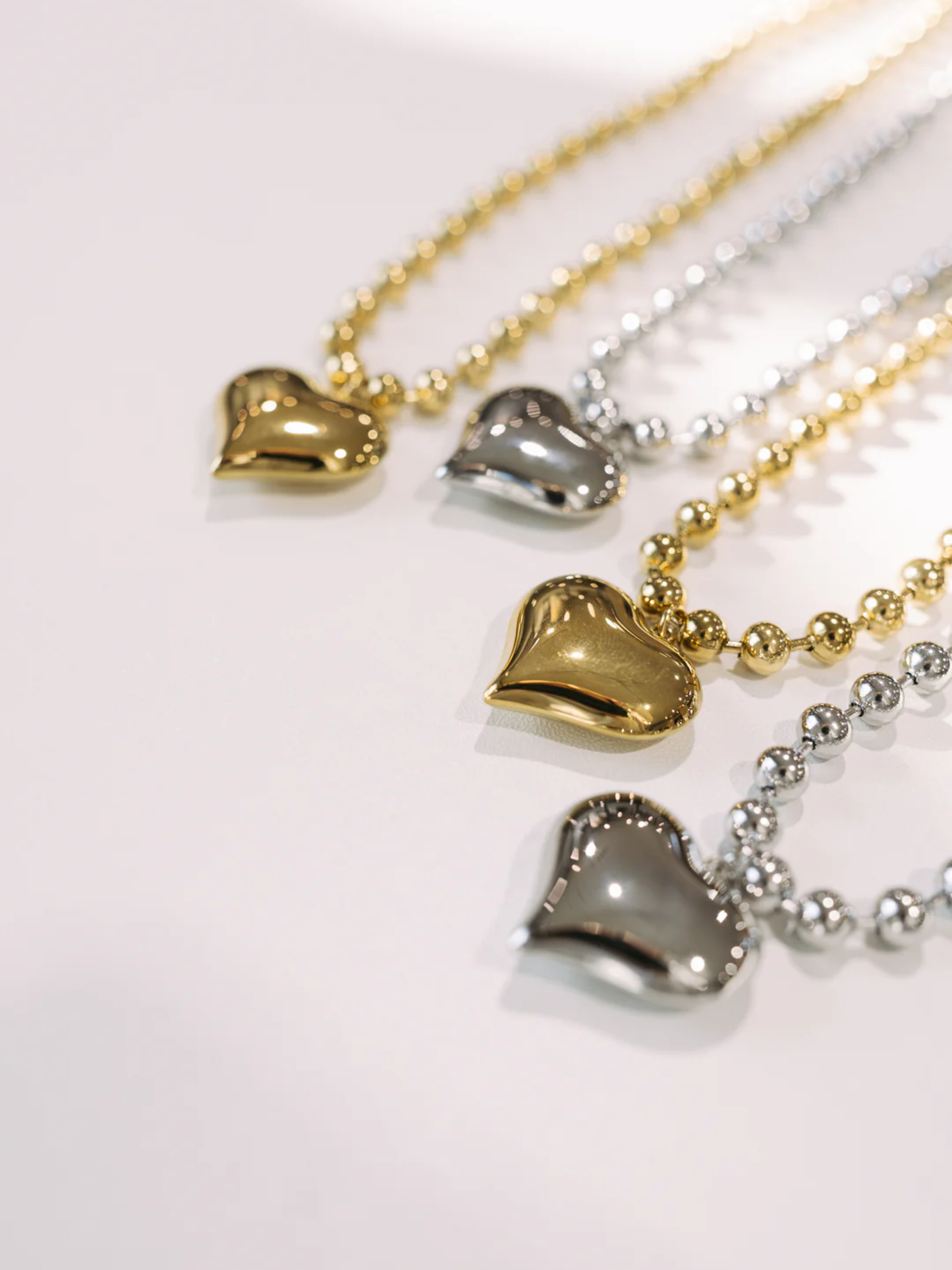 Four puffy heart pendant beaded necklaces in gold and silver tones on white surface.